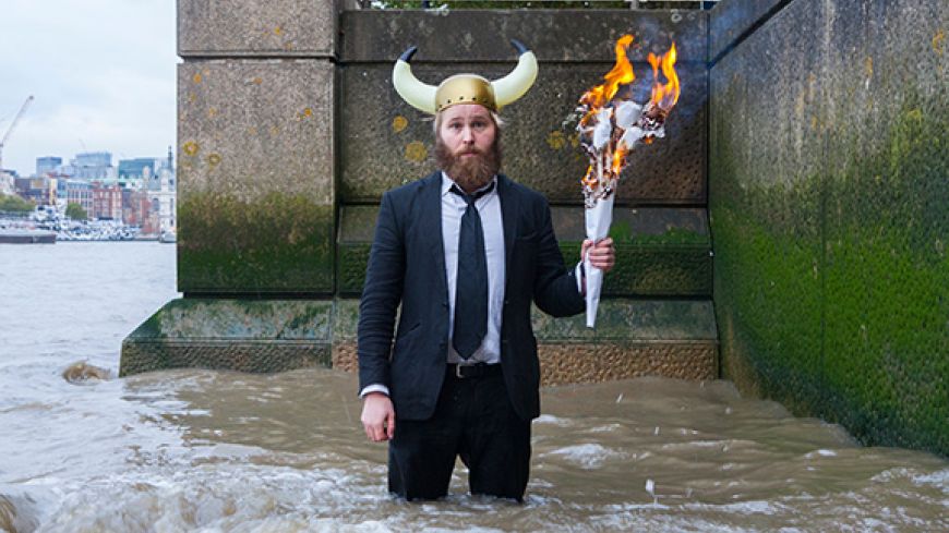 James Rowland, dressed in a suit with a viking helmet on his head, stands in the river