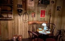 A darkened room with a table, chairs, and wooden walls. Teapot and glasses on the table.