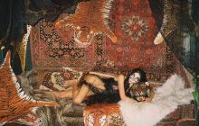 A bikini clad woman wearing joke glasses, lies on a bed of faux tiger skins and Persian rugs, draped in a long black hair wig