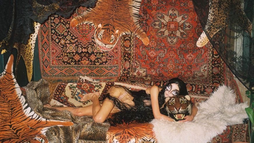 A bikini clad woman wearing joke glasses, lies on a bed of faux tiger skins and Persian rugs, draped in a long black hair wig