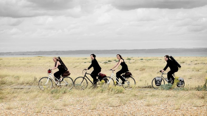 Four musicians ride through a field with instruments on their backs