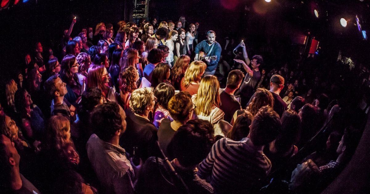 A room full of people singing, black background, with two performers in the middle.