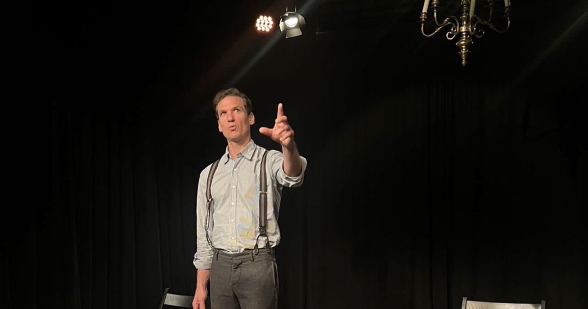 Actor Luke Townson stands looking out, with a background of a black box theatre space, chairs and books visible