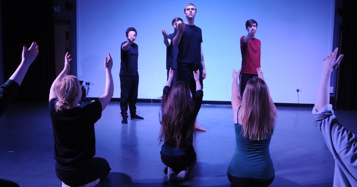 A white and blue lit backdrop highlights the outline of performers in lines, arms reaching up, from the show Death by Shakesepeare