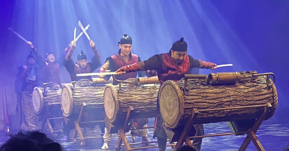 Drummers in a line, lit by blue light, with white beams highlighting