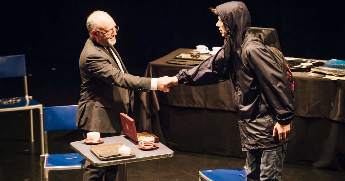 Two men greet each other with a handshake. One is dressed in a suit, the other in a rain coat with a hood.