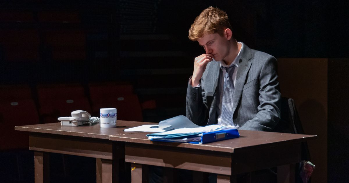 The lead actor as The Lawyer, sits at a table looking at papers, with a thoughtful look on his face.