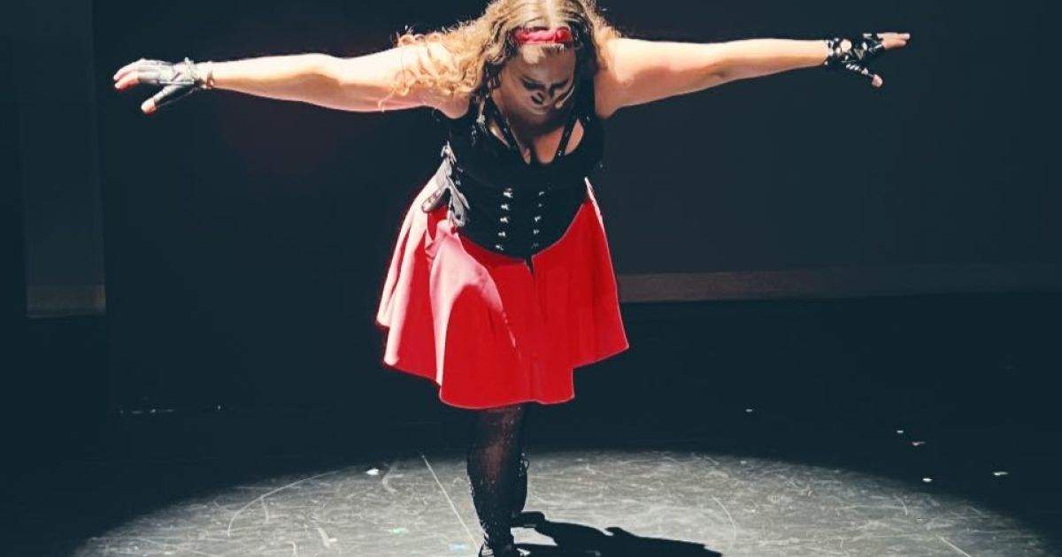 Performer Torrey Shine, dressed in black bustier, pink skirt, looks down with her arms outstretched, lit in a circle, but surrounded by darkness