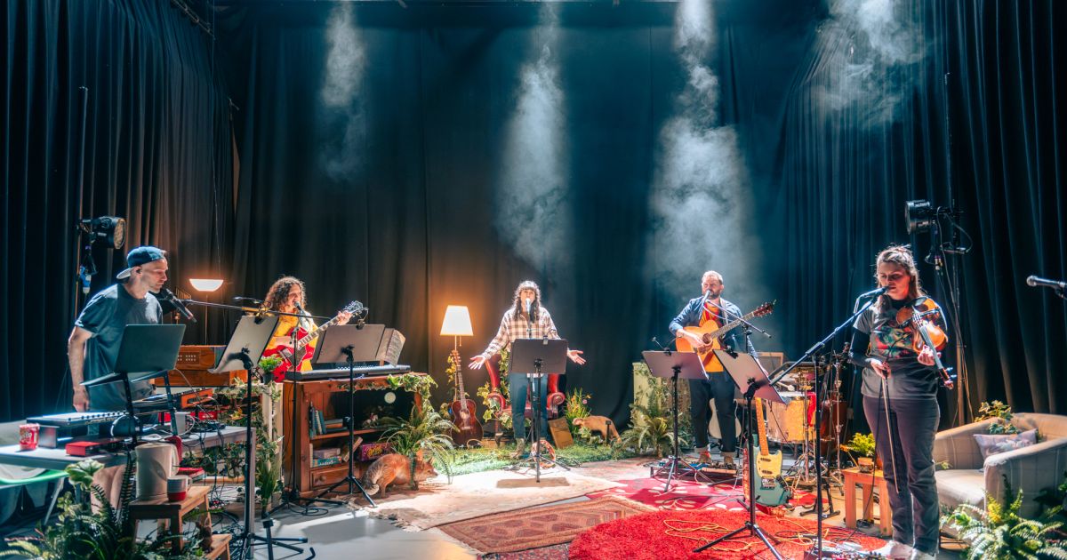 Five performers are light in a smoky atmosphere, with greenery, in a half circle