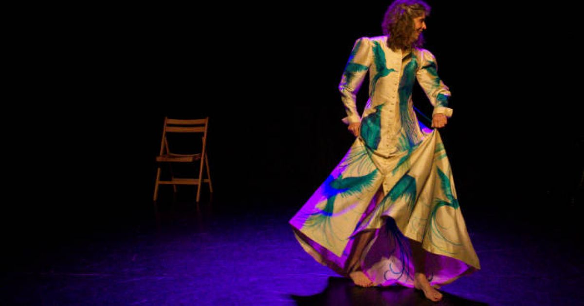 Performer Maria MacDonell starts, highlighted in a dark background, in a long glamorous cream coloured robe that has large bright blue peacocks on it.