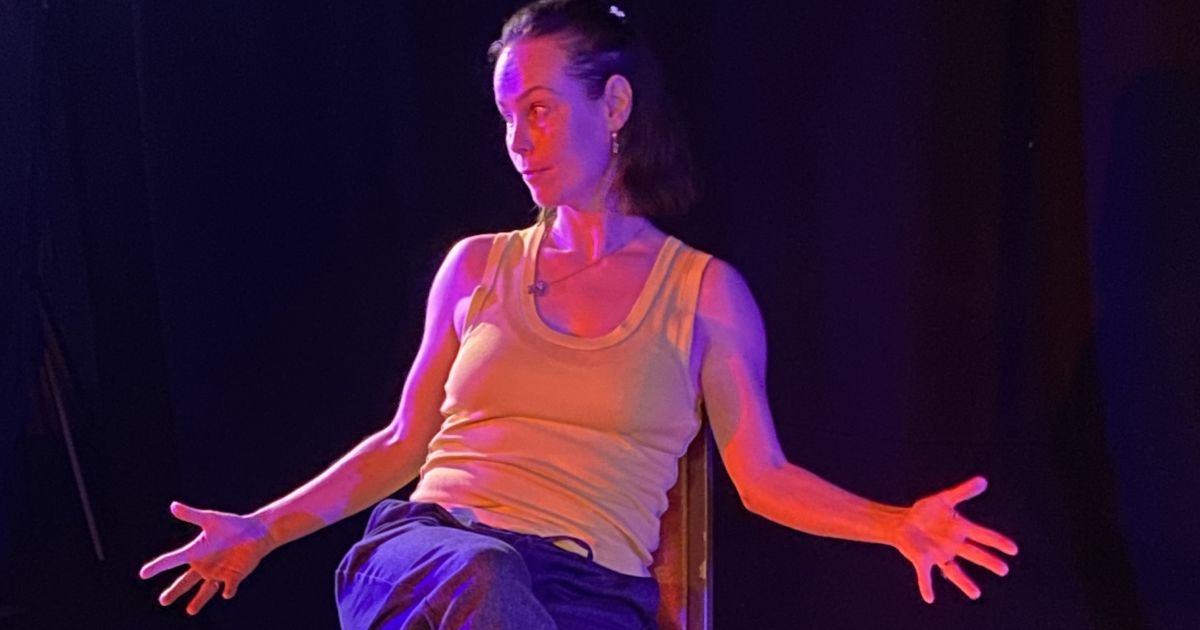 Performer Ambrosine Davies sits in a chair, with her arms extended, in dark light with a spotlight. She wears dark blue jogging trousers and a lime green vest.