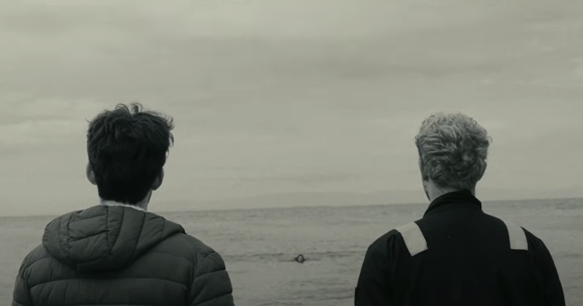 Two young men stand and look out at the wide open sea. The day seems grey and gloomy.