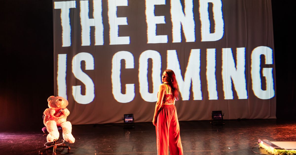Screen with the words 'the end is coming' angled across it. Performance artist Louise Orwin stands in front of it with her back to the audience, wearing a red strappy top and red tracksuit bottoms. A giant teddy bear is seated to the left of the screen.