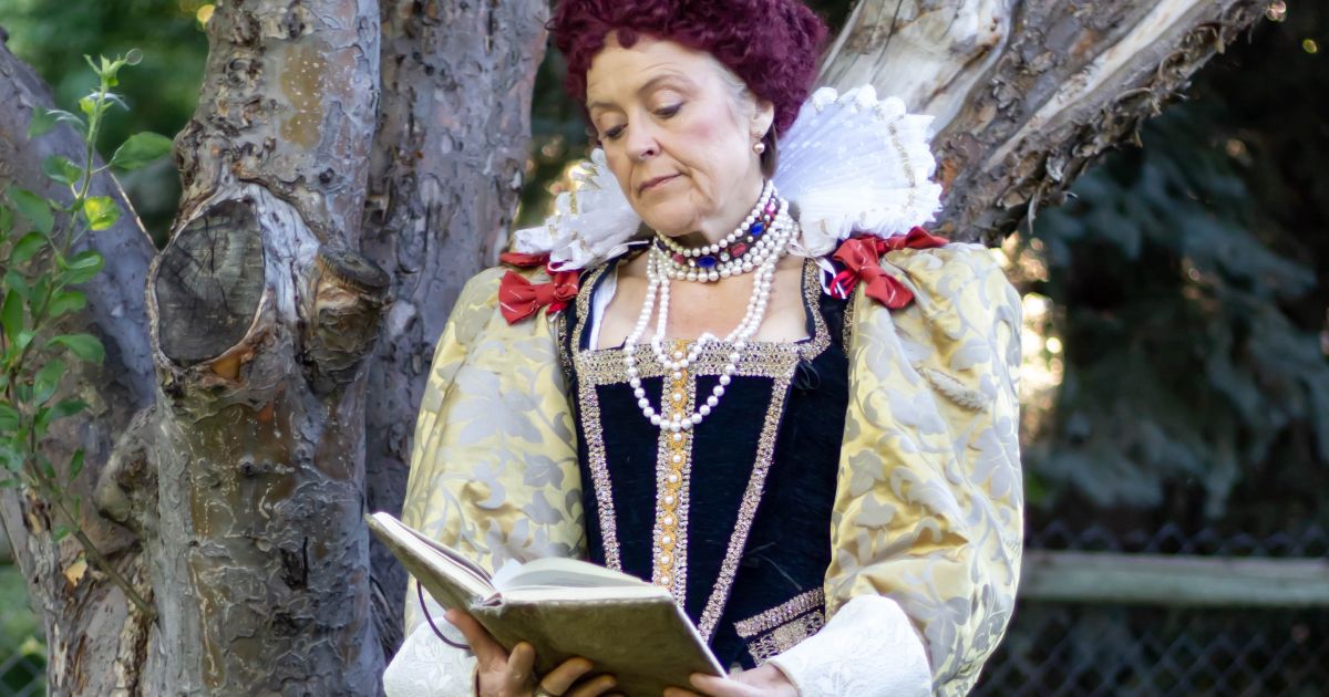 Elizabeth in her wig and full costume, looking at a book with her head tilted.