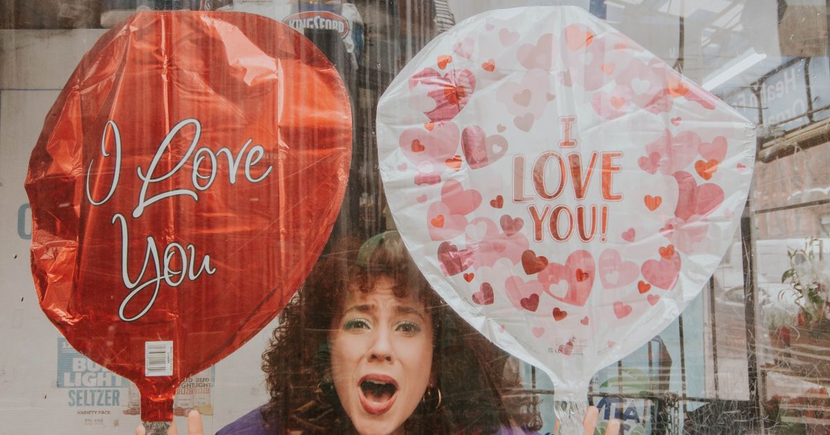 Writer/Performer Gianna Milici holds two balloons, one red, one pink. She wears a long green scarf around her neck.