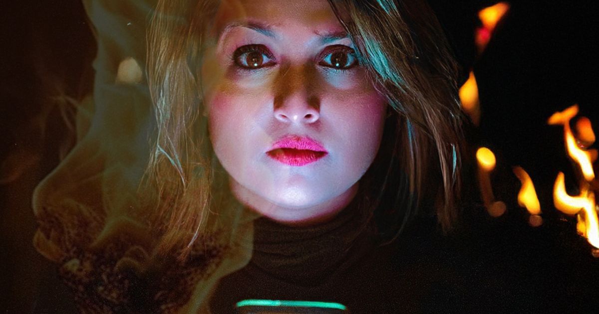 Woman looks directly at the screen. Darkness is behind with some fire. Fierce look.