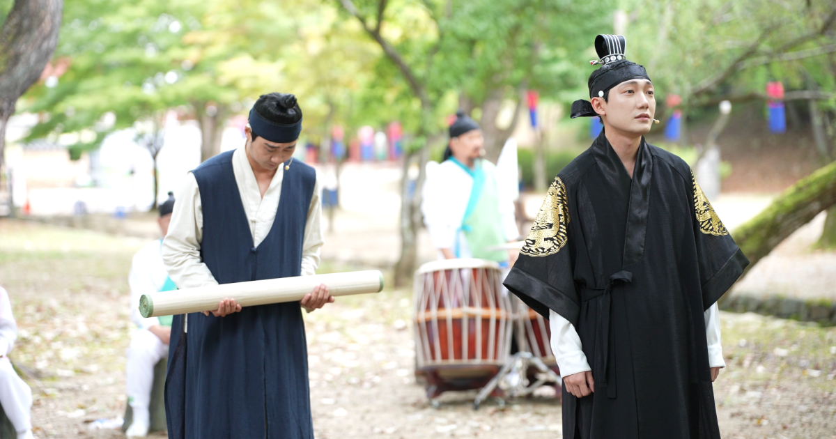 A courtier stands to the left of the King, with the Queen in the background, pictured in a green leafy area.