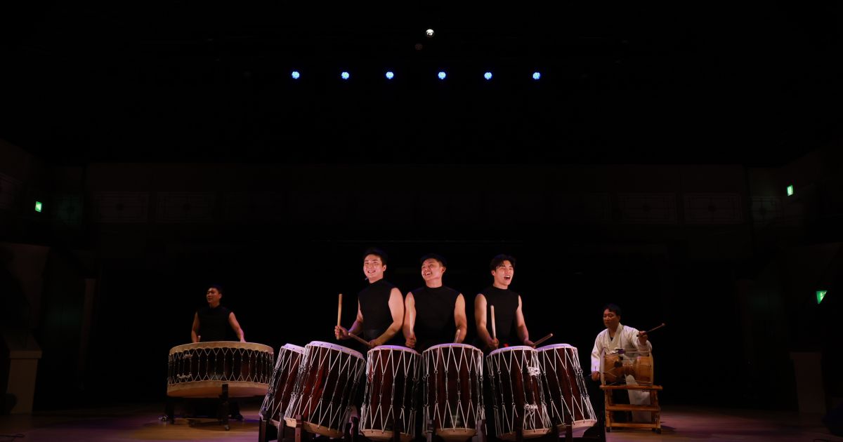 Drummers are pictured with their drums, in front of a dark background.