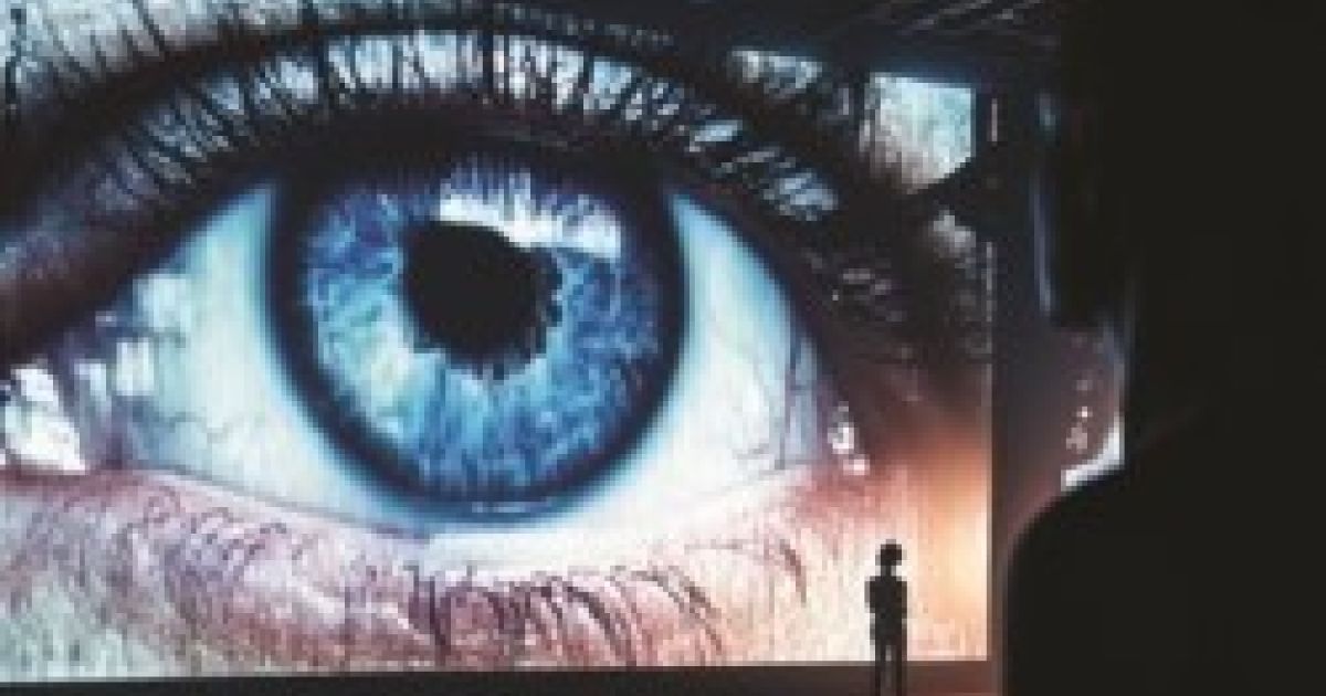 An eye watches over you. A person stands in front of it, very small in comparison.