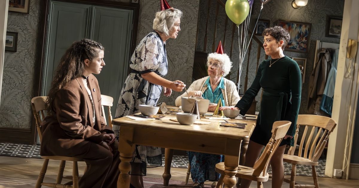 The four actors in the play are pictured round a kitchen table, all looking at each other, balloons in the background.