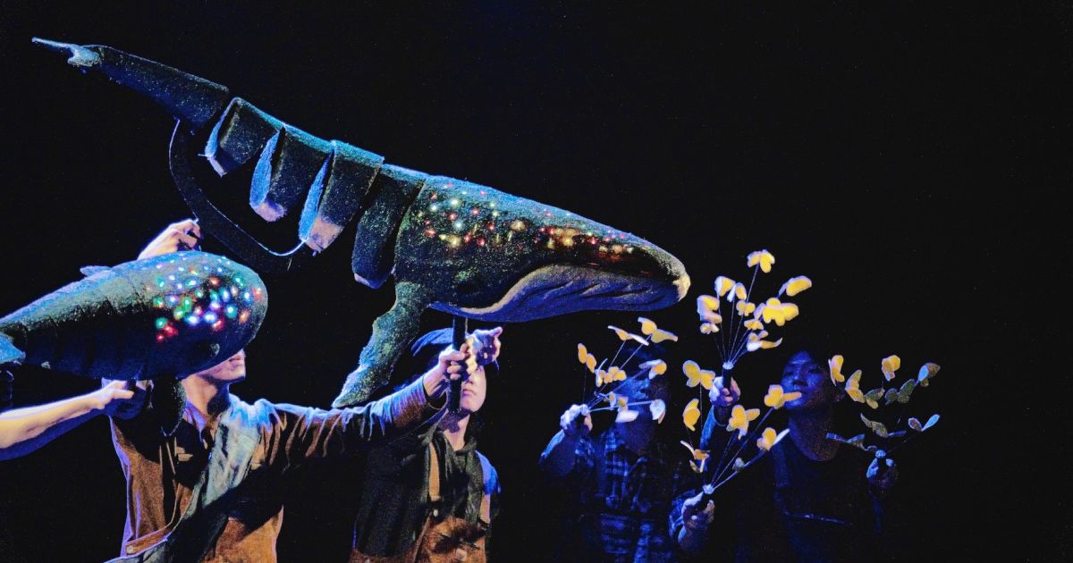 Whale puppet moves, held by puppeteers. Dark background, whale is lit up with lights showing on head.