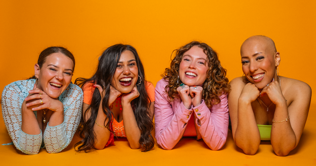 The four members of the girl group lie on the ground, with their faces on their crossed arms. Background is a mustard yellow. All are smiling.