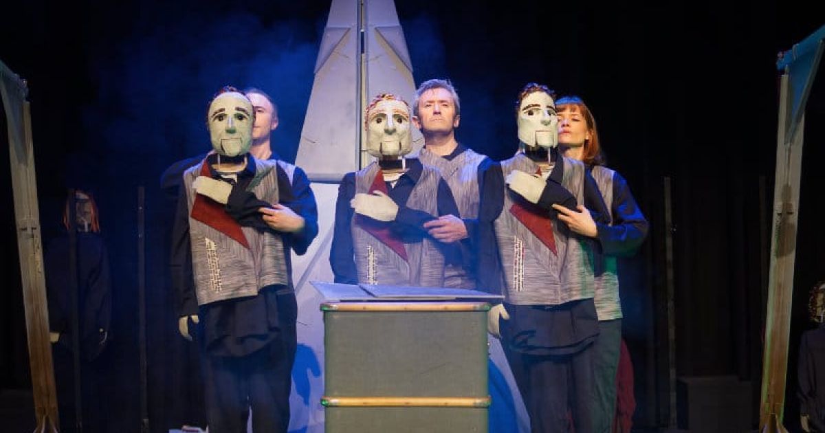 Performers on stage with the puppet masks, stand in the centre, large item at the back behind them