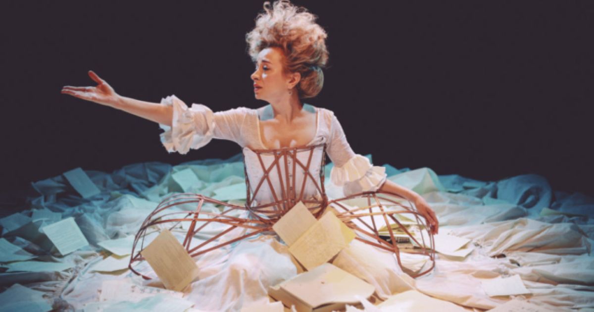 A woman sits on the ground, surrounded by a big white dress. Her hair is piled high and messy and one arm is outstretched.