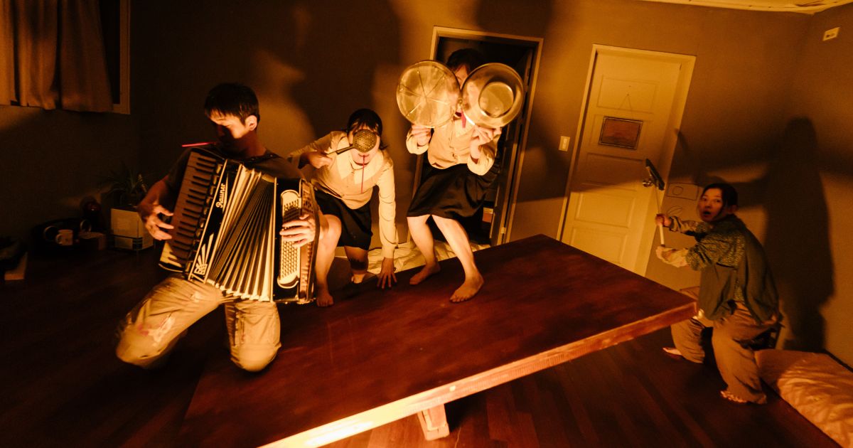 A musican plays; 3 other actors hide behind objects in a dark, shadowy room.