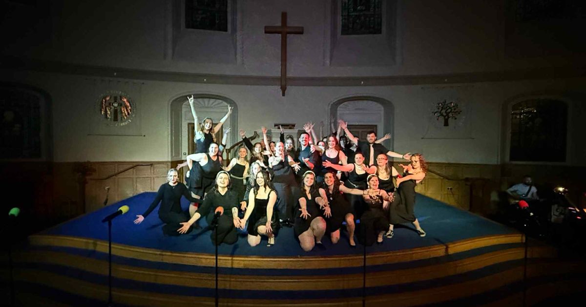 Large cast photo pose with arms out, black background, all seen in the spotlight. Church background shown.