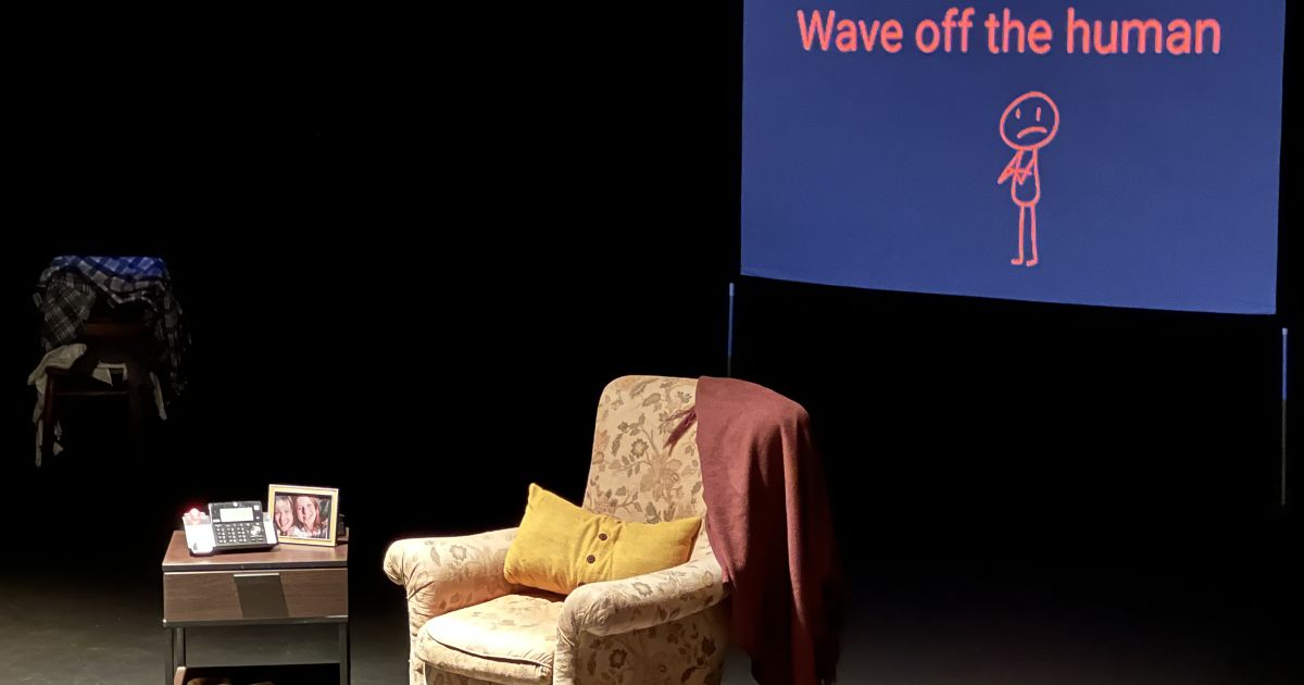 A screen shows in the background, an armchair and small bedside table to the side. The lighting is dark, with the furniture lit.