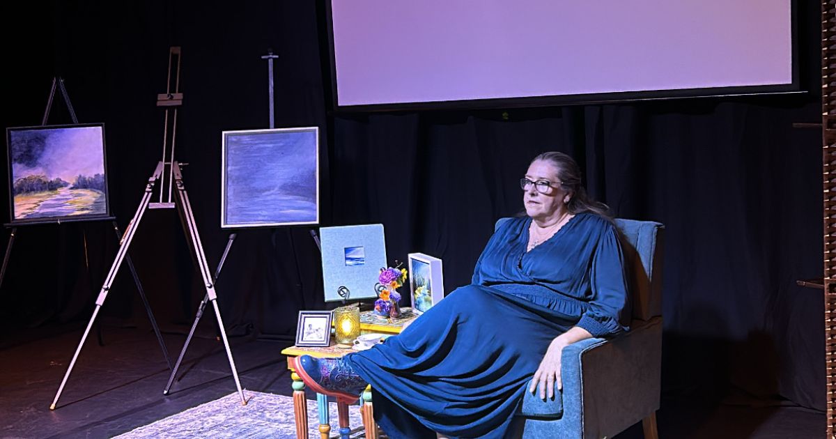 Performer/playwright Patricia McGrath sits next to a screen. There is dark blue light and paintings to the side. Everything is awash with shades of blue.