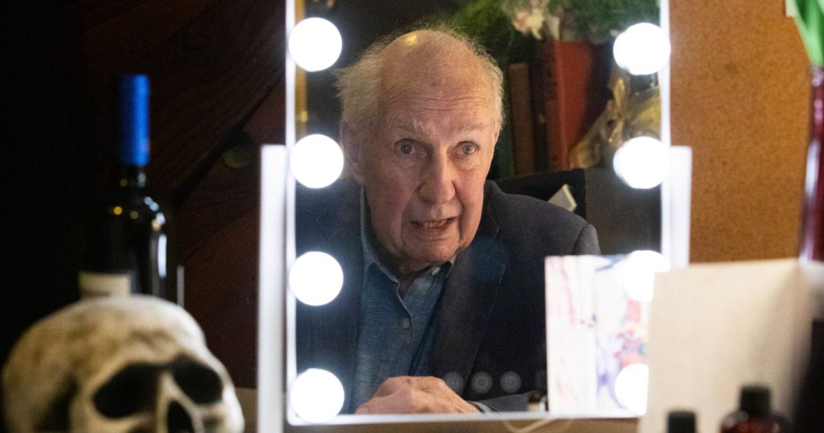 Nick Ullett, the actor and writer, is reflected in a dressing room mirror, lights around it, and a skull to his left.