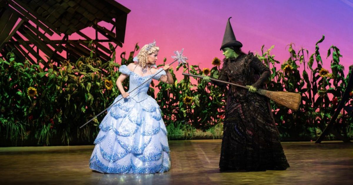 Sarah O'Connor and Laura Pick (Glinda and Elphaba) face off with brooms. Glinda wears a sparkly blue ballgown, Elphaba her traditional witch's outfit and has her green body colouring. 