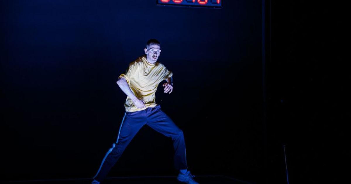 A male dancer is lit in the centre of a dark space with a screen showing numbers in red in the background