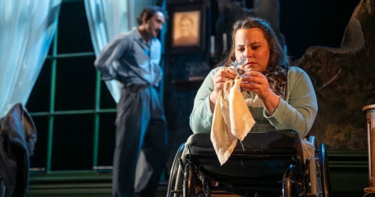 Amy Conachan as Laura sits polishing a glass figure as Christopher Jordan-Marshall as Tom hovers in the background arguing offstage with their mother.