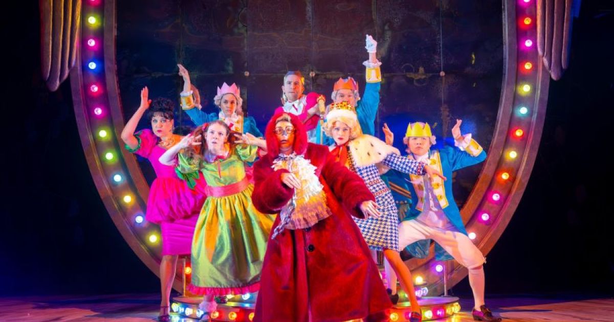 The full cast of Cinderella A Fairytale pose in front of a giast crest, dressed in very bright clothing.
