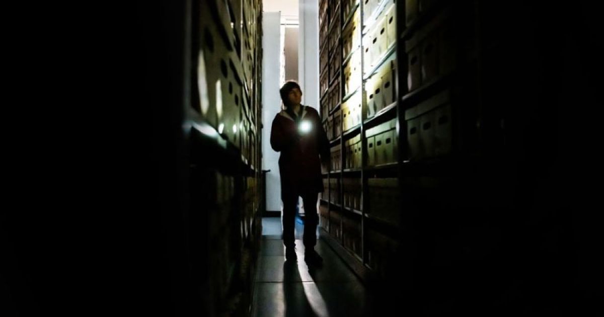 Danny Robins. strides between shelving, covered in box files, moody lighting in black and white