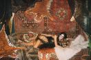 A bikini clad woman wearing joke glasses, lies on a bed of faux tiger skins and Persian rugs, draped in a long black hair wig